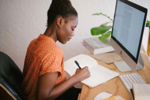 A young woman writes in a notebook while reading on her computer.