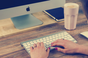 The hands of a woman shown typing on a computer keyboard with a coffee drink nearby.