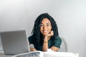 A young woman with glasses smiles while sitting behind a laptop. 