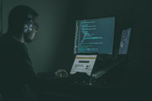 A man in a dark room does coding on dual computer monitors.