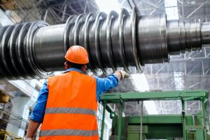 A man in an orange safety vest and hardhat examines an advanced manufacturing process on a factory floor.