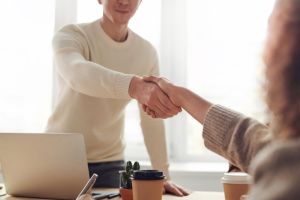 A man and a woman shake hands over a desk with a laptop on it. 