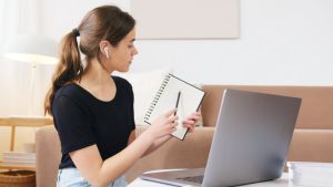 A young woman with long brown hair wearing a black shirt gestures to a writing pad while speaking on a video call. 