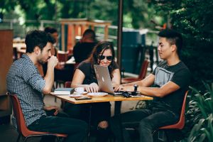 Three college students sitting at a table with one of them working on a laptop